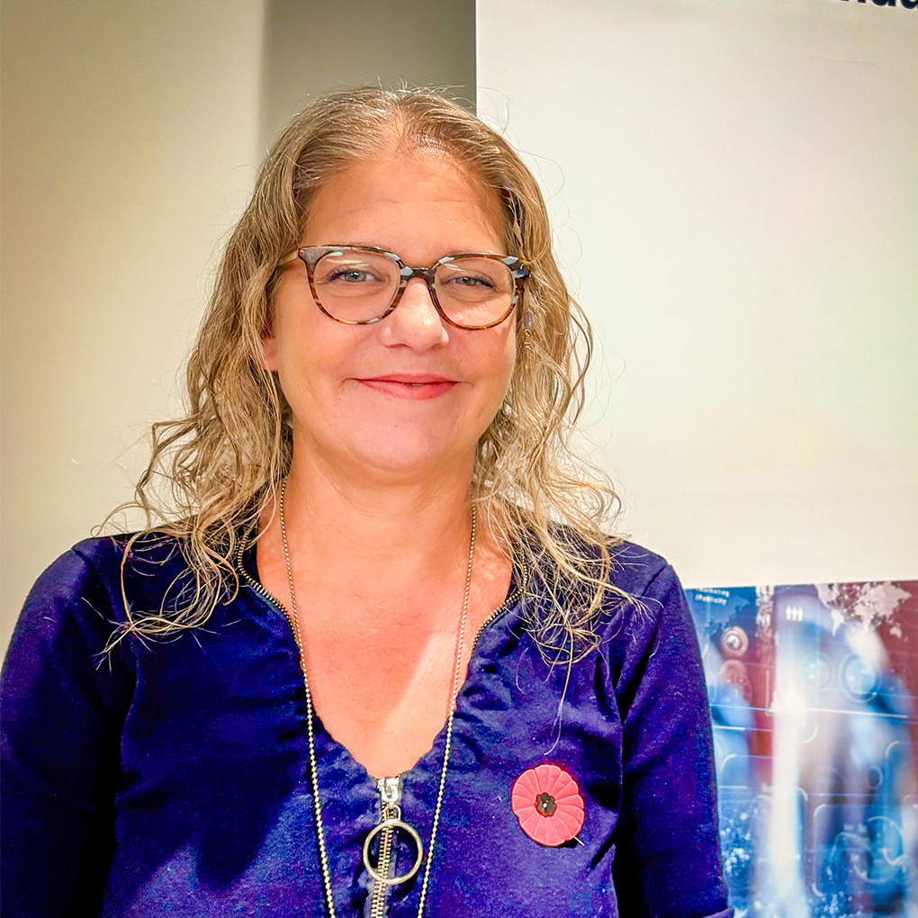 Woman wearing glasses and a purple top with a red poppy, smiling in a studio setting.