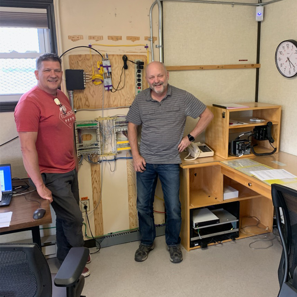 Two men standing in a workshop beside shelving and technology equipment, smiling at the camera.