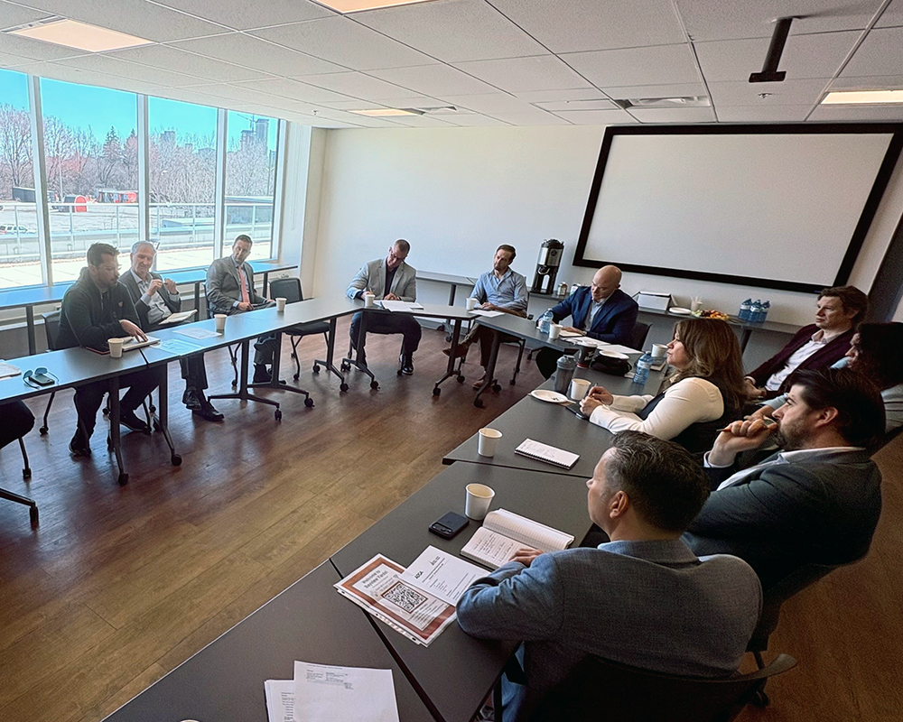 A group of people in business attire are seated around tables in a conference room, engaged in a meeting. Large windows let in natural light, and a projector screen is visible in the background.