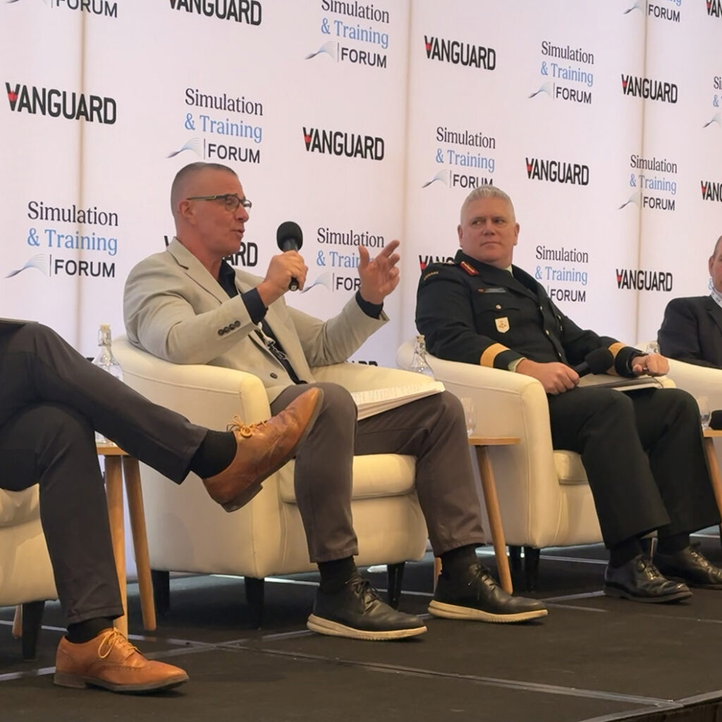 Four men sit on stage in armchairs during a panel discussion. One man, holding a microphone, gestures while speaking. Another man in a military uniform sits beside him. Banners behind them read "Vanguard" and "Simulation & Training Forum".