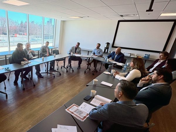 A group of people in business attire are seated around tables in a conference room, engaged in a meeting. Large windows let in natural light, and a projector screen is visible in the background.
