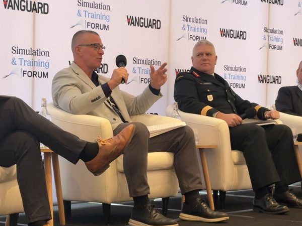 Four men sit on stage in armchairs during a panel discussion. One man, holding a microphone, gestures while speaking. Another man in a military uniform sits beside him. Banners behind them read "Vanguard" and "Simulation & Training Forum".